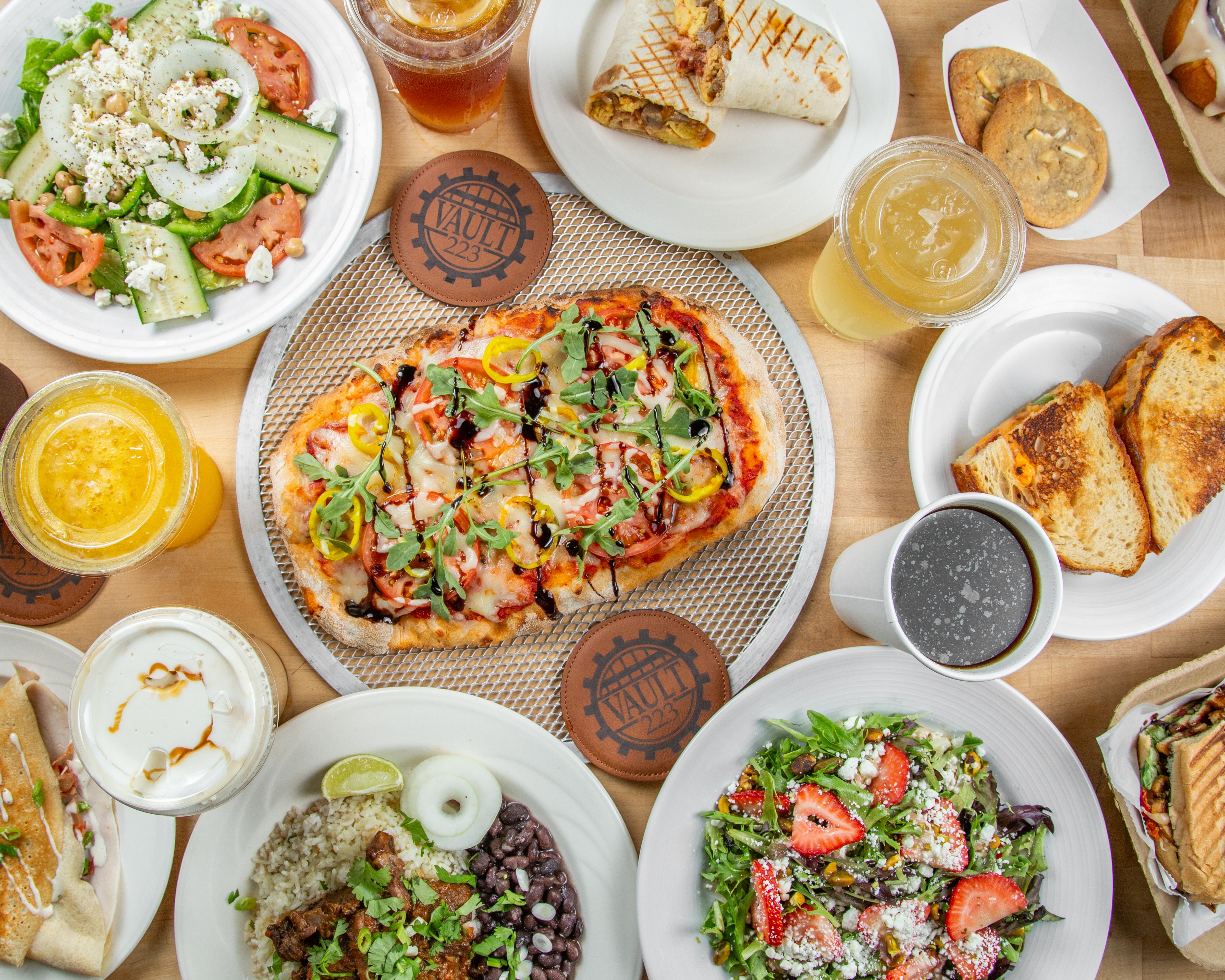 An overhead spread of Vault 223 pizza, salads, breakfast, sandwiches, crepes, drinks, and branded coasters